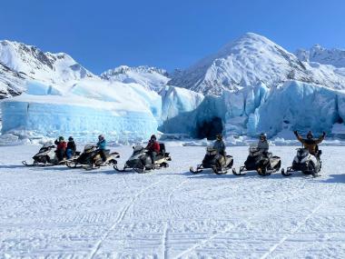 Snowmobilers pose in front of a glacier on a snowmobile tour.