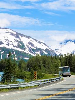 A motorcoach drives an Alaska roadway beside a river and mountains