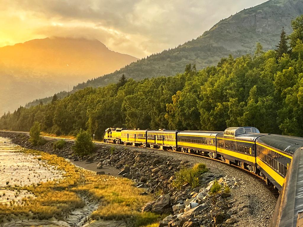 A yellow train travels along a scenic stretch of Alaska Railroad's Costal Classic route as the sun begins to set